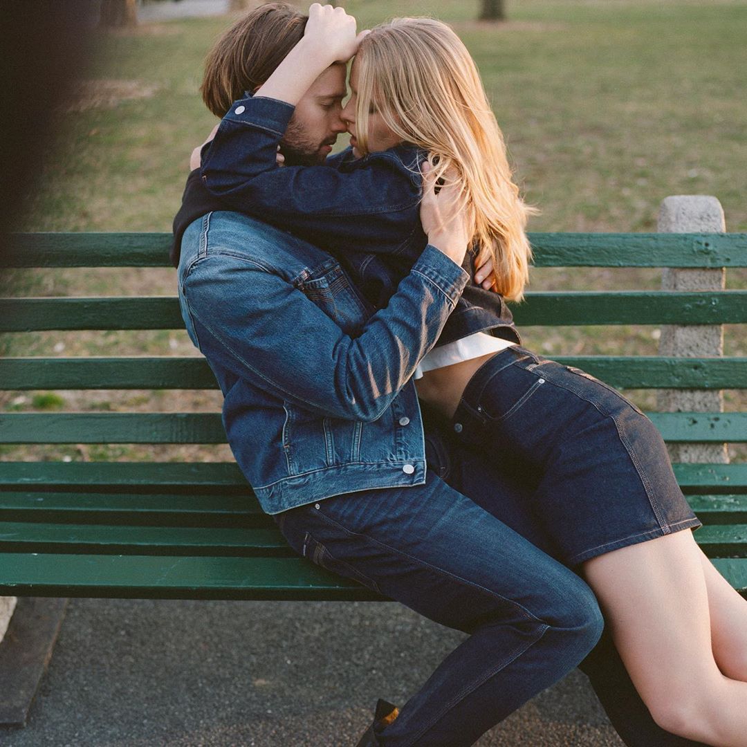 A close-up shot of a couple embracing on a green park bench, dressed in denim jackets and jeans, with a blurred nature background.