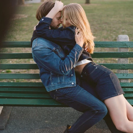 A close-up shot of a couple embracing on a green park bench, dressed in denim jackets and jeans, with a blurred nature background.