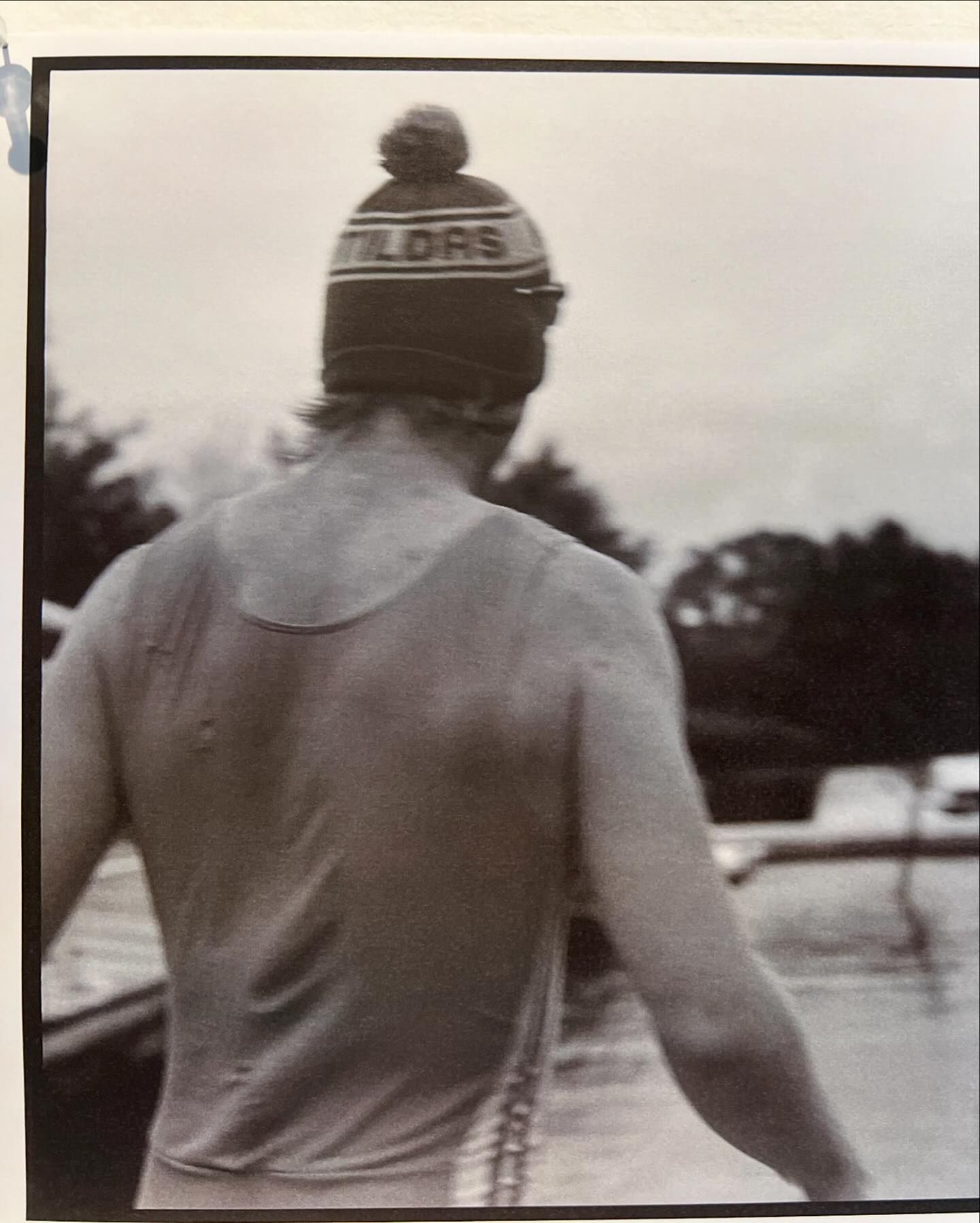 A person faces away from the camera, wearing a tank top and a knit cap with the text 'TILDES' on it, near a body of water.