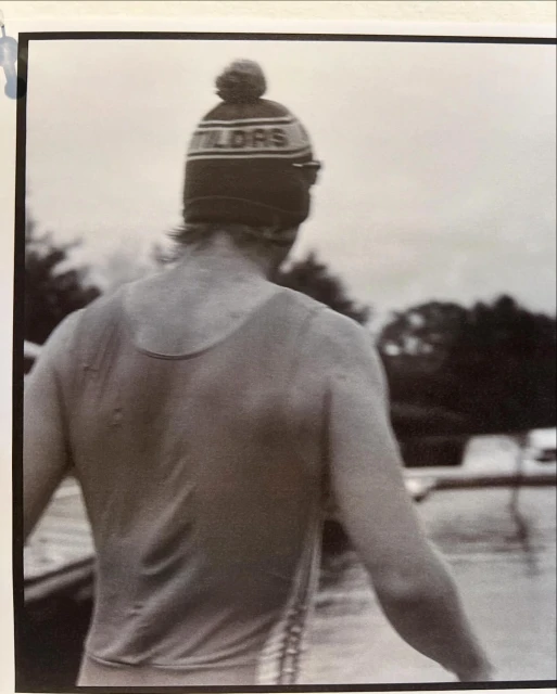 A person faces away from the camera, wearing a tank top and a knit cap with the text 'TILDES' on it, near a body of water.