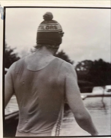 A person faces away from the camera, wearing a tank top and a knit cap with the text 'TILDES' on it, near a body of water.