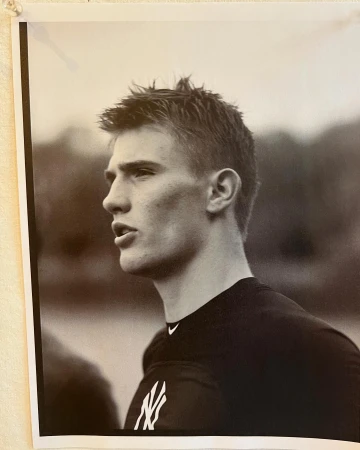 Monochrome portrait of a young man wearing a t-shirt with a logo, photographed in profile against an outdoor backdrop.