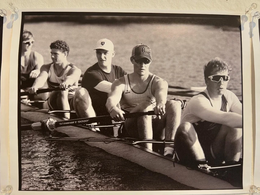 Monochrome image of a rowing team in action on the water, featuring several young men in a racing shell, oars in motion, creating ripples.