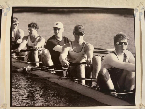 Monochrome image of a rowing team in action on the water, featuring several young men in a racing shell, oars in motion, creating ripples.