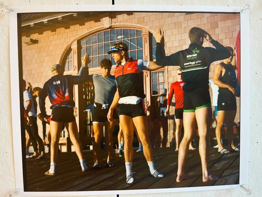 Several athletes in rowing team apparel stretch their arms up while standing on a deck near a building with an arched window.
