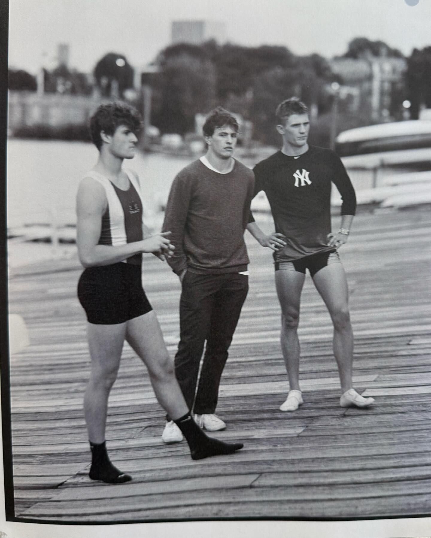 Three young adult males stand on a wooden pier wearing athletic outfits in a black-and-white photo; city buildings in background.