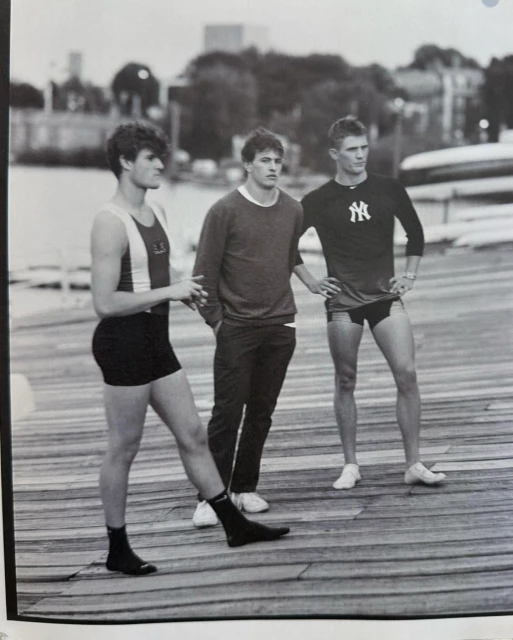 Three young adult males stand on a wooden pier wearing athletic outfits in a black-and-white photo; city buildings in background.