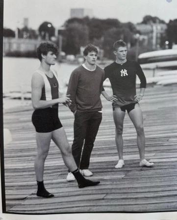 Three young adult males stand on a wooden pier wearing athletic outfits in a black-and-white photo; city buildings in background.