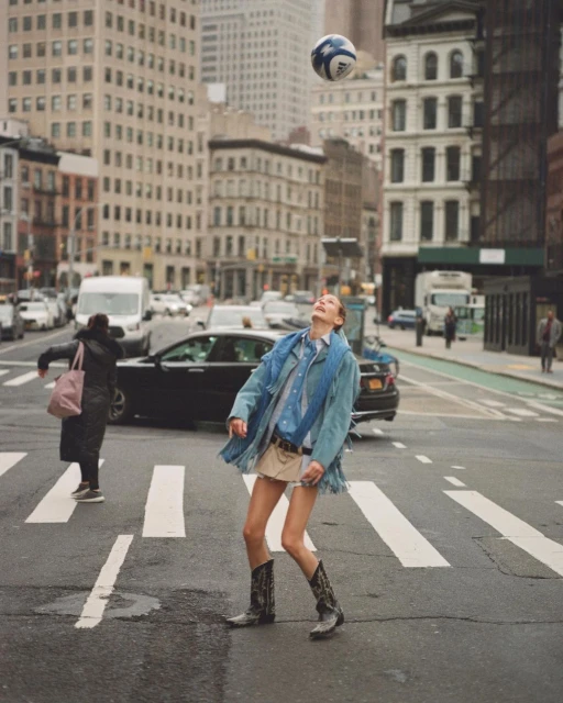 A woman in cowboy boots and denim jacket plays with a soccer ball on a crosswalk in a city with tall buildings in the background.