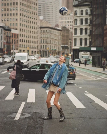 A woman in cowboy boots and denim jacket plays with a soccer ball on a crosswalk in a city with tall buildings in the background.