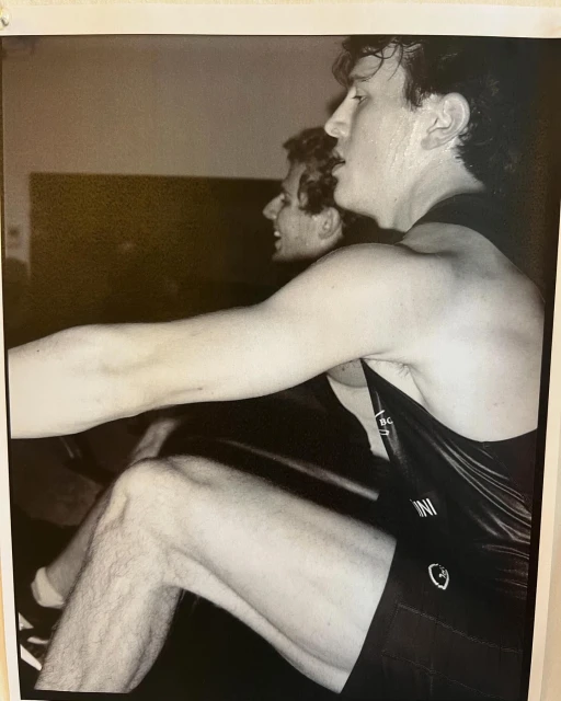 Monochrome photo of two adult males rowing indoors, focused on the foreground rower's profile, arms, and legs, wearing a tank top and shorts.