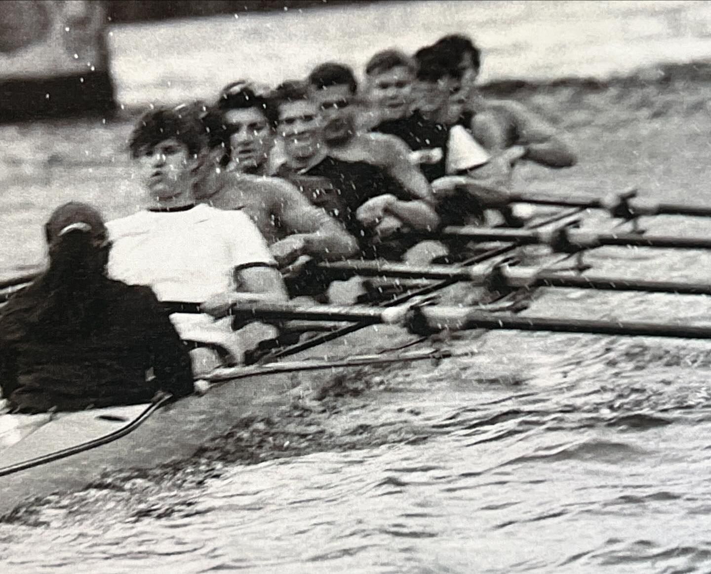 Eight young adult rowers compete in a crew boat race on choppy water in a monochrome, eye-level action shot.