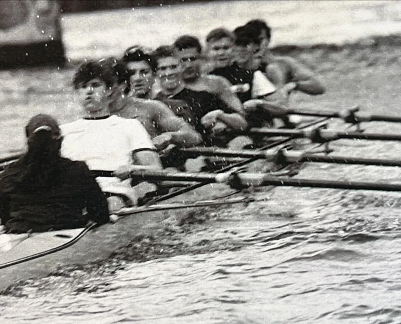Eight young adult rowers compete in a crew boat race on choppy water in a monochrome, eye-level action shot.