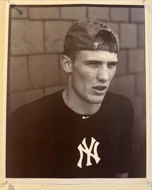 Black and white portrait of a young adult male wearing a baseball cap and New York Yankees t-shirt, posing in front of a brick wall.