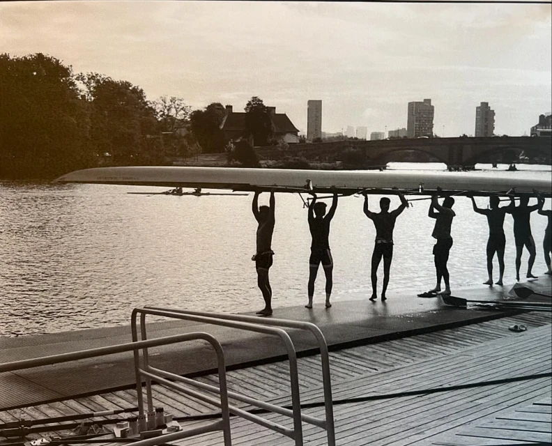 Rowing team carries a scull over their heads on a dock along a river with buildings in the background on an overcast day.