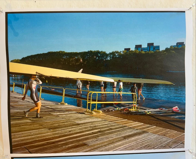 Rowing team carries a yellow shell on a wooden dock by the water. A woman in a grey rowing uniform walks in the foreground.