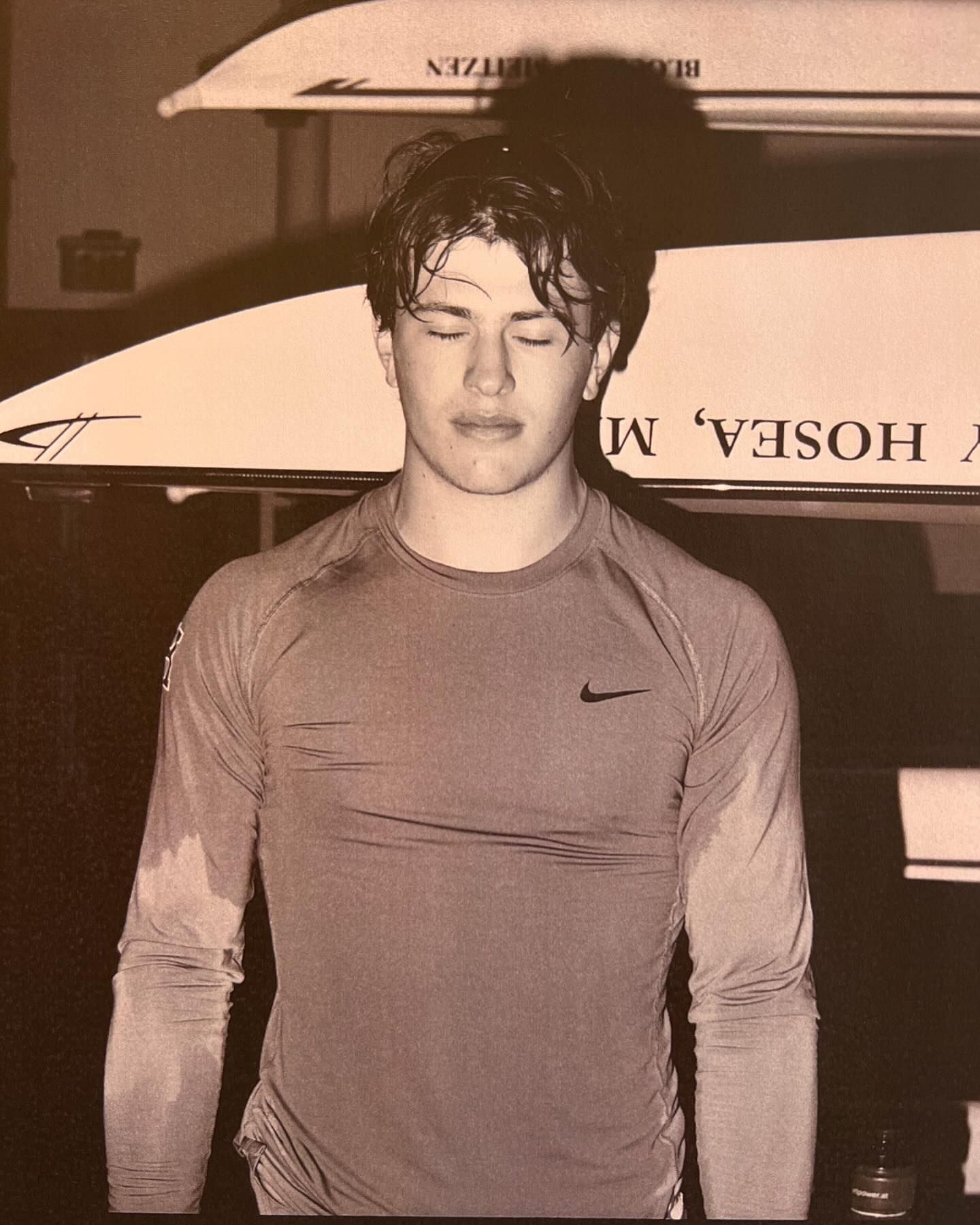 Monochromatic portrait of a wet, athletic man with his eyes closed, framed by rowing sculls above in an indoor environment.