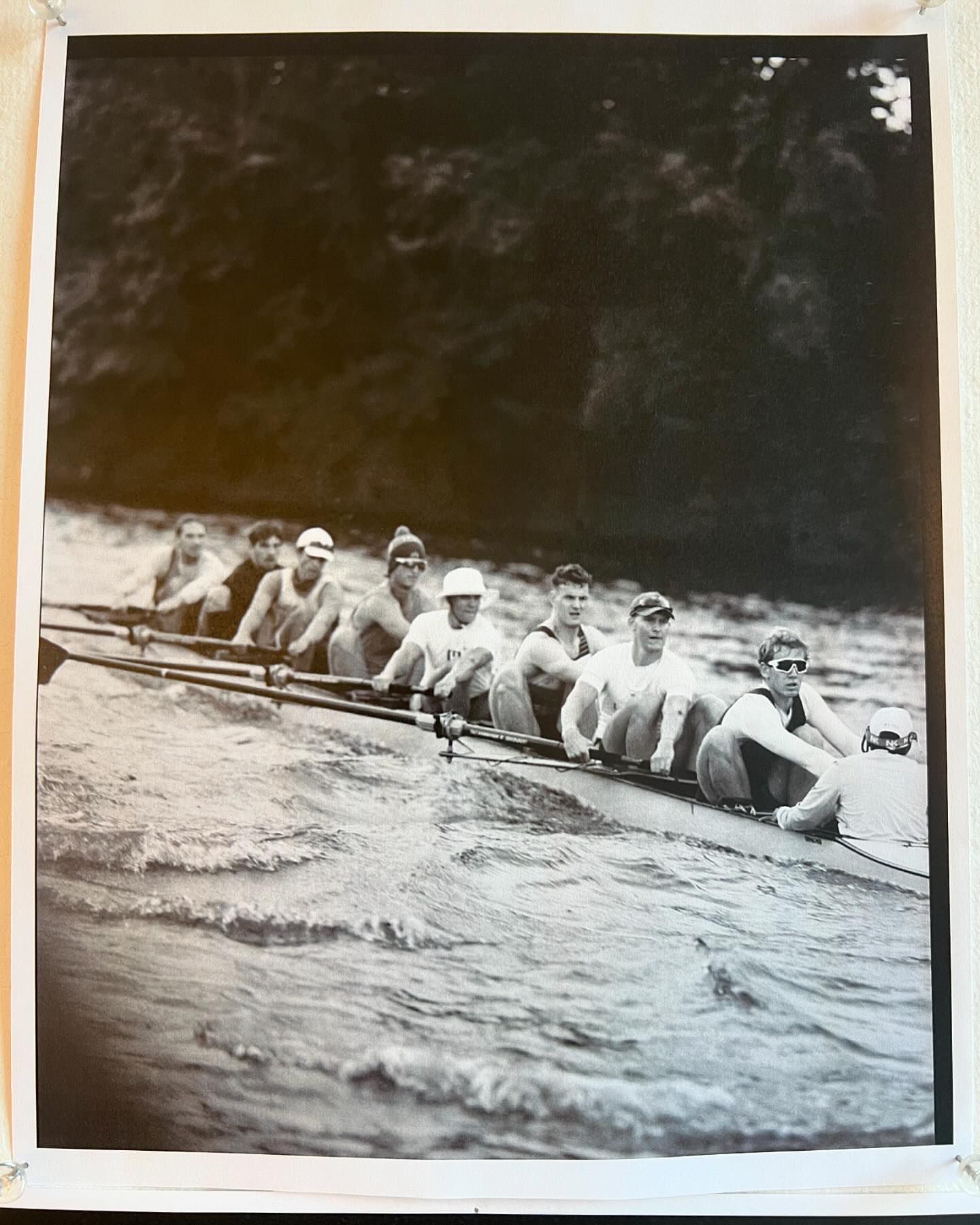 Black and white photo of a rowing team in action on a wavy body of water with trees visible in the background, capturing the sport's intensity.
