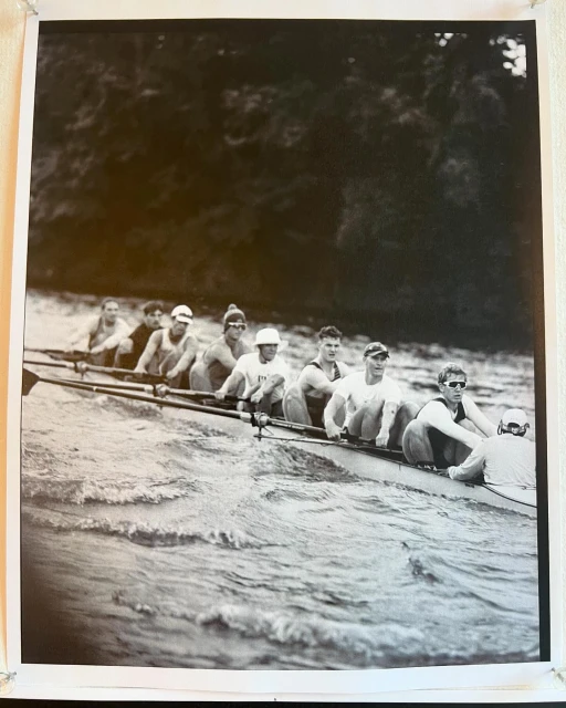 Black and white photo of a rowing team in action on a wavy body of water with trees visible in the background, capturing the sport's intensity.