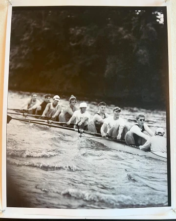 Black and white photo of a rowing team in action on a wavy body of water with trees visible in the background, capturing the sport's intensity.