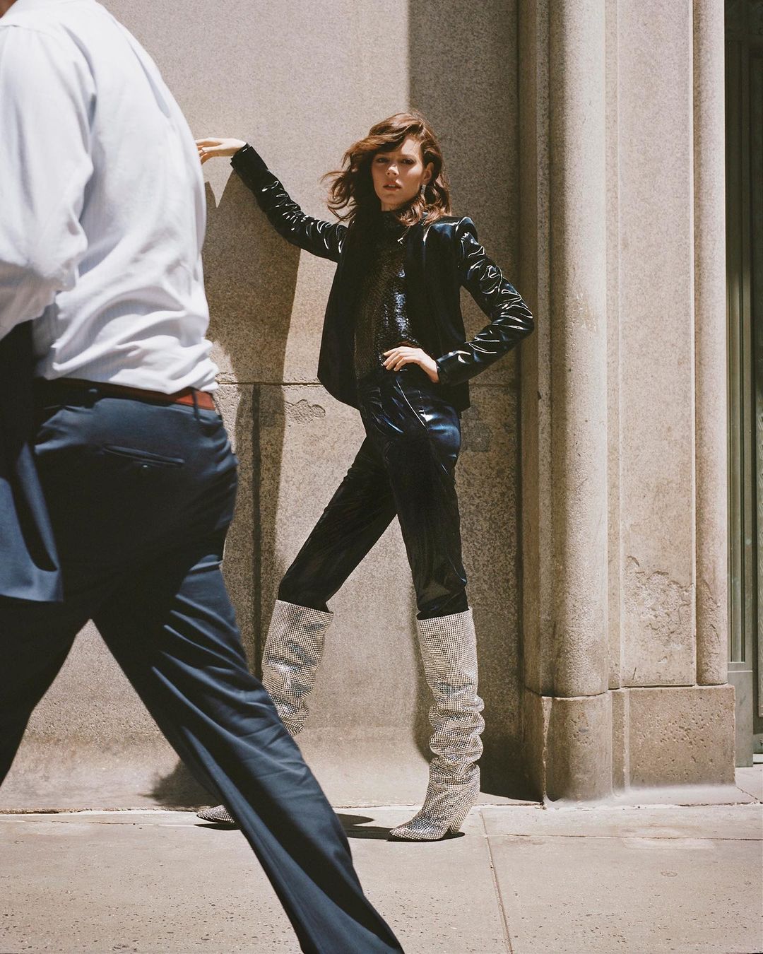 Fashionable woman in leather jacket and boots poses against stone wall as blurred figure passes by on city street during daylight.