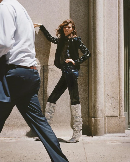 Fashionable woman in leather jacket and boots poses against stone wall as blurred figure passes by on city street during daylight.