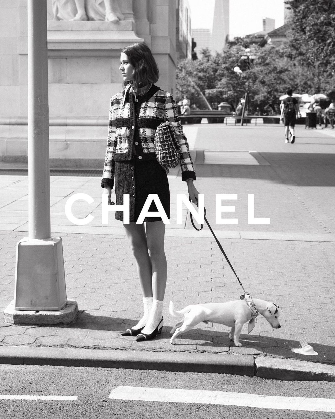 Black and white outdoor shot of a woman in Chanel fashion posing on a city sidewalk, walking a Jack Russell Terrier dog on a leash.