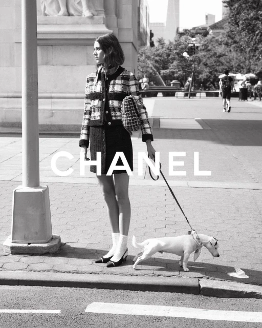 Black and white outdoor shot of a woman in Chanel fashion posing on a city sidewalk, walking a Jack Russell Terrier dog on a leash.