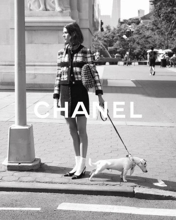 Black and white outdoor shot of a woman in Chanel fashion posing on a city sidewalk, walking a Jack Russell Terrier dog on a leash.