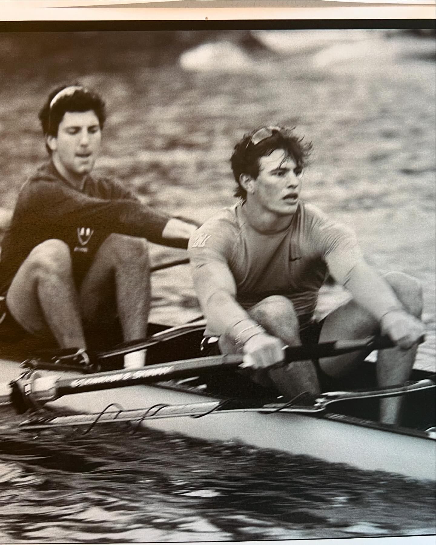 Two adult males rowing a scull on a body of water in a black and white photo showcasing teamwork and physical exertion.