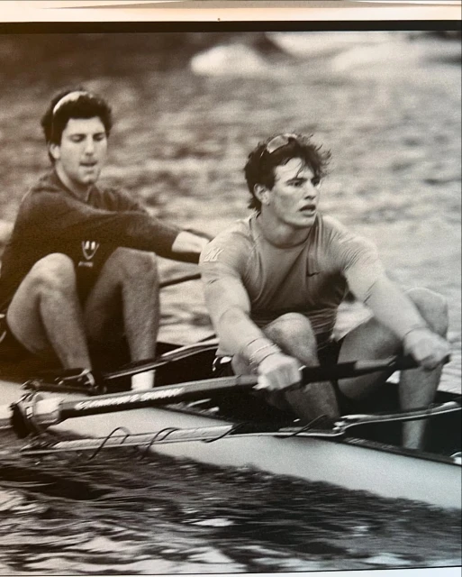 Two adult males rowing a scull on a body of water in a black and white photo showcasing teamwork and physical exertion.