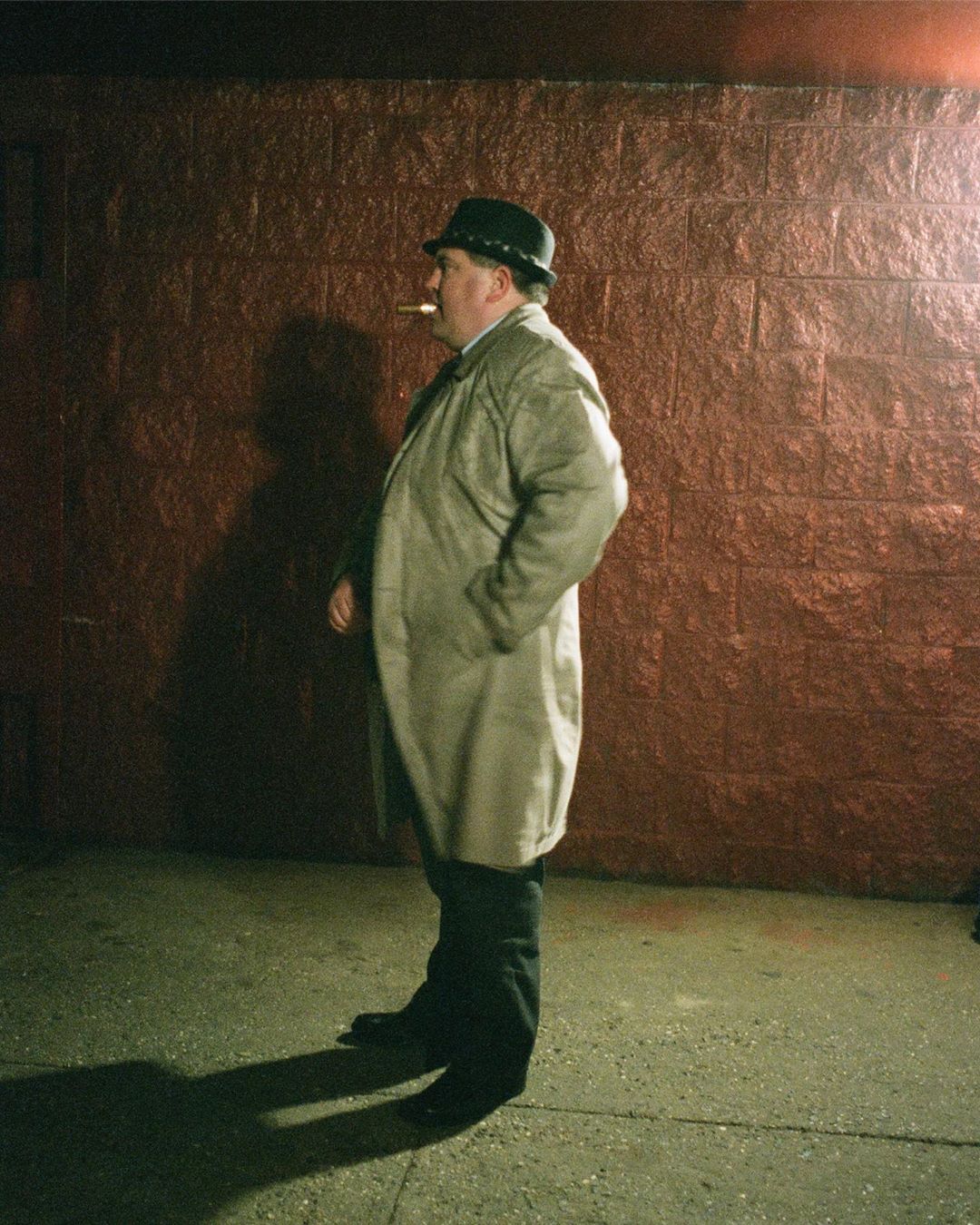 Man in coat and hat smoking against a brick wall in an outdoor urban setting, illuminated by a street light.