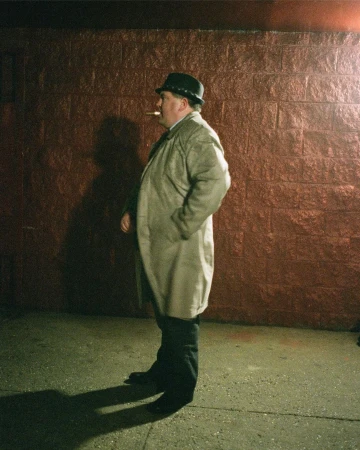 Man wearing a fedora hat and trench coat holding a cigar while standing against a red brick wall at night