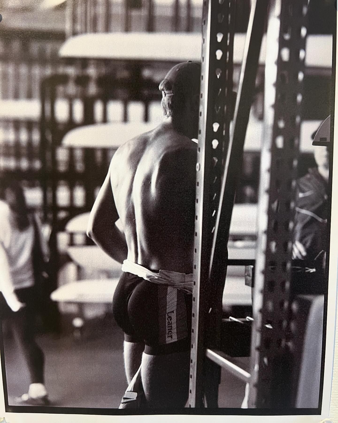 Black and white shot from behind of muscular man with a cap, wearing swim trunks with the word “Leaner” on the side, between metal shelves.