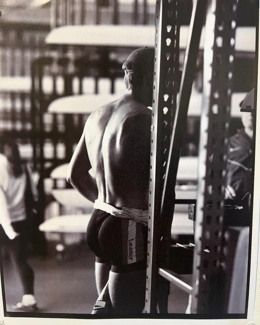 Black and white shot from behind of muscular man with a cap, wearing swim trunks with the word “Leaner” on the side, between metal shelves.