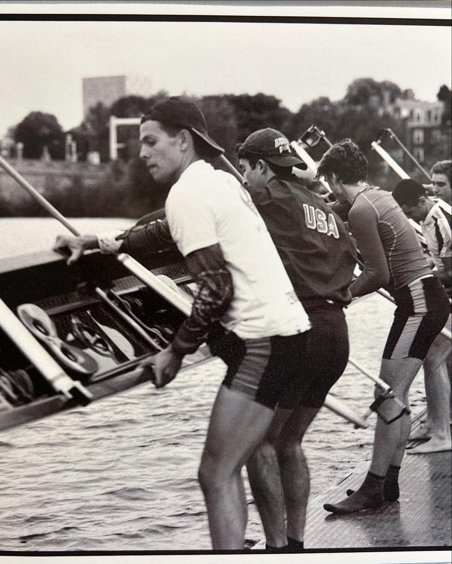 Monochrome image of a rowing team carrying a shell on a river, athletes in sportswear, with buildings and trees visible in the background.