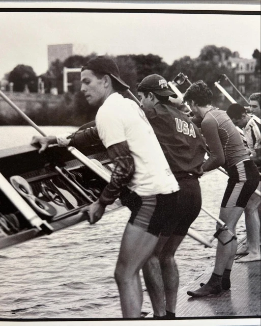Monochrome image of a rowing team carrying a shell on a river, athletes in sportswear, with buildings and trees visible in the background.