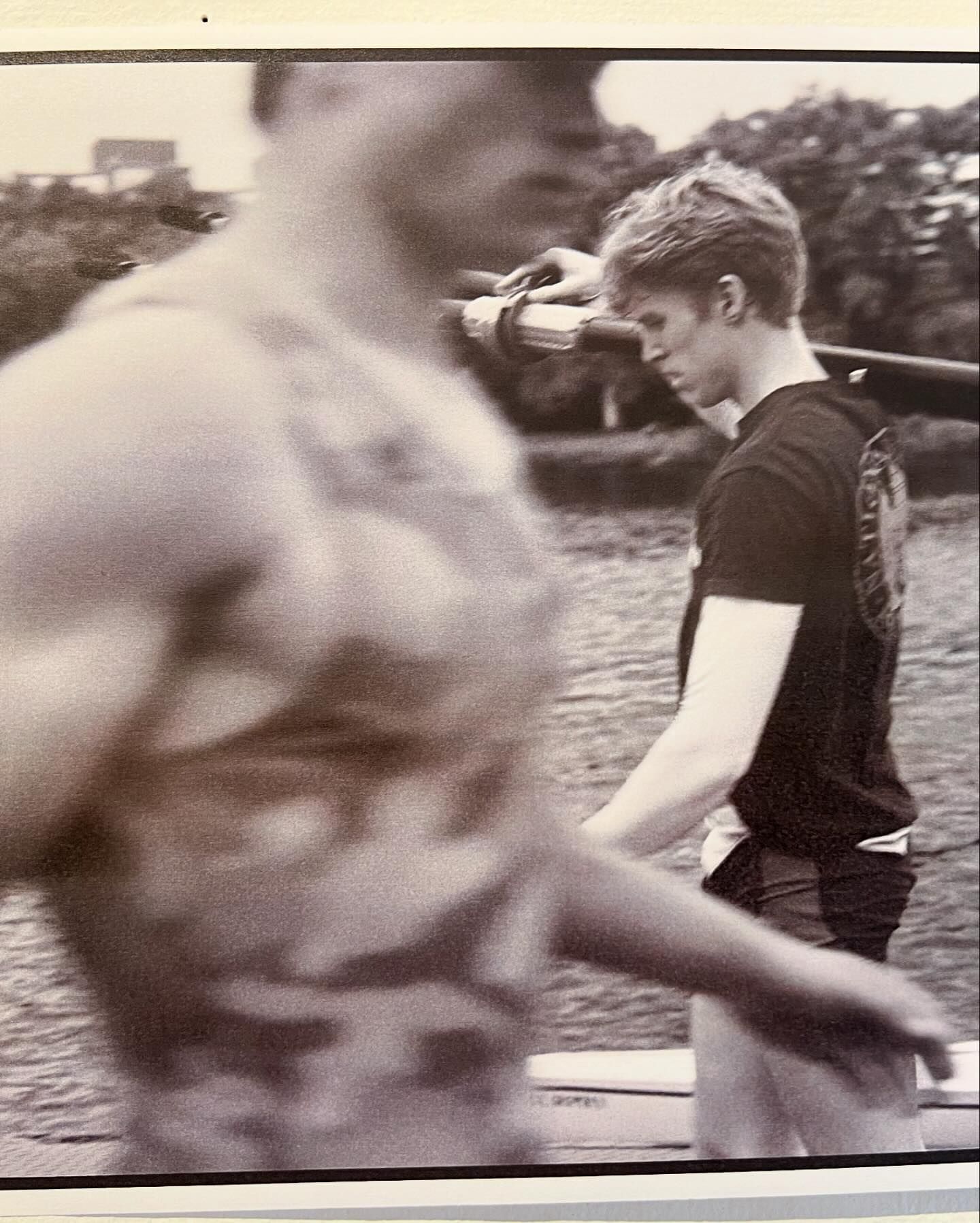 Two men carry a rowing shell outdoors. One man, in focus, wears a black shirt and the other, blurred, wears a patterned tank top.