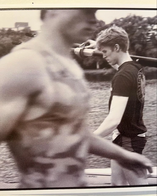 Two men carry a rowing shell outdoors. One man, in focus, wears a black shirt and the other, blurred, wears a patterned tank top.