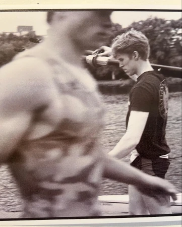 Two men carry a rowing shell outdoors. One man, in focus, wears a black shirt and the other, blurred, wears a patterned tank top.