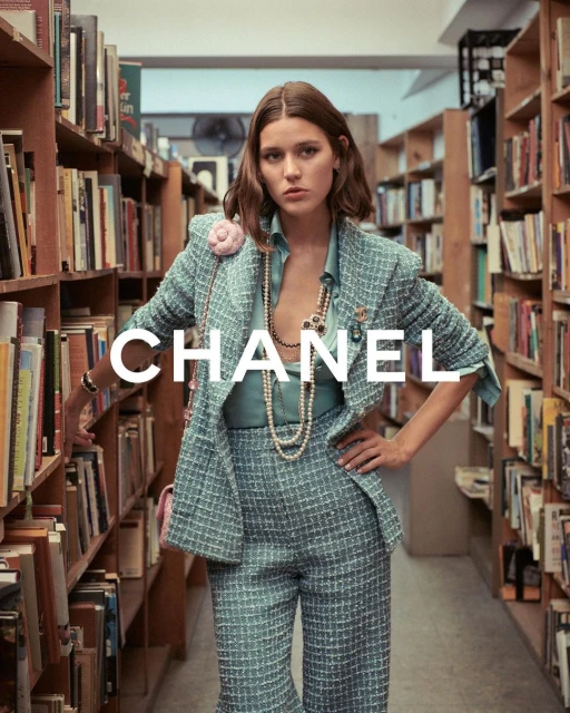Fashion shot of a young woman in a tweed suit and jewelry posing in a library with bookshelves and the text overlay 'CHANEL'.