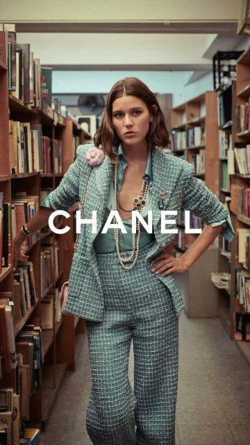 Fashion shot of a woman in a teal pantsuit standing in a library with rows of books, styled with camellia brooch, pearl necklaces, and wrist jewelry.