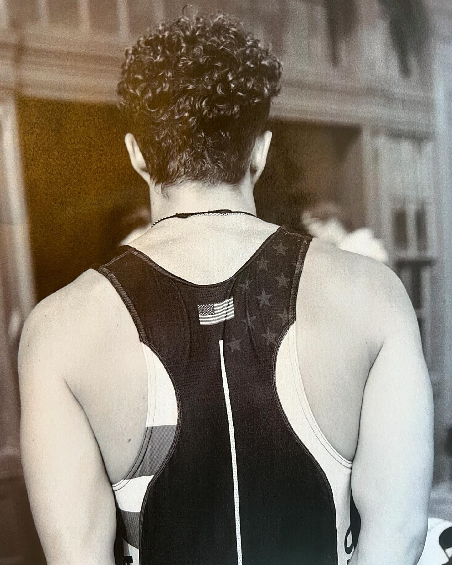 Back view of an adult male in a singlet with the American flag on it. He has curly hair and is shot in black and white.