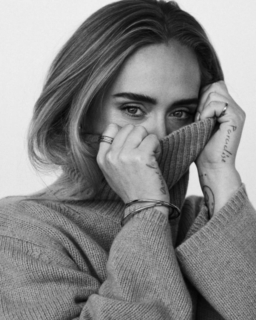 Close-up portrait of a woman with blonde hair wearing a turtleneck sweater, her hands partially covering her face, in black and white.