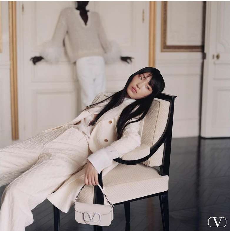 Fashion shot of an Asian woman in a cream suit, holding a Valentino bag, posing in a chair, and a black model stands in the background.