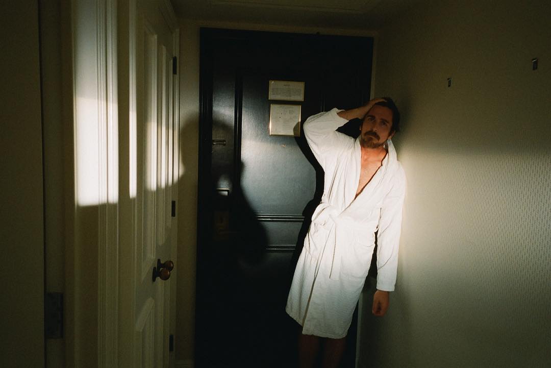 A man with a beard wearing a white robe, standing in a room with a dark door and neutral walls, lit by window light.