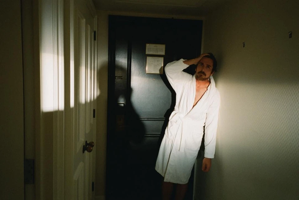 A man with a beard wearing a white robe, standing in a room with a dark door and neutral walls, lit by window light.