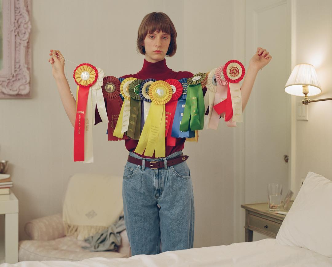 Portrait of a young adult woman indoors with neutral expression, wearing a turtleneck and jeans, draped in various award ribbons.