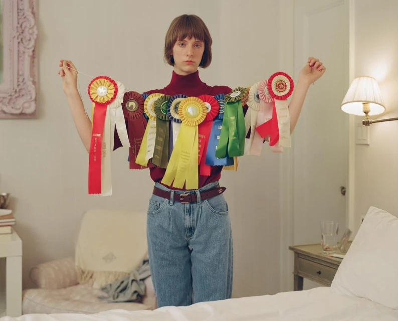 Portrait of a young adult woman indoors with neutral expression, wearing a turtleneck and jeans, draped in various award ribbons.