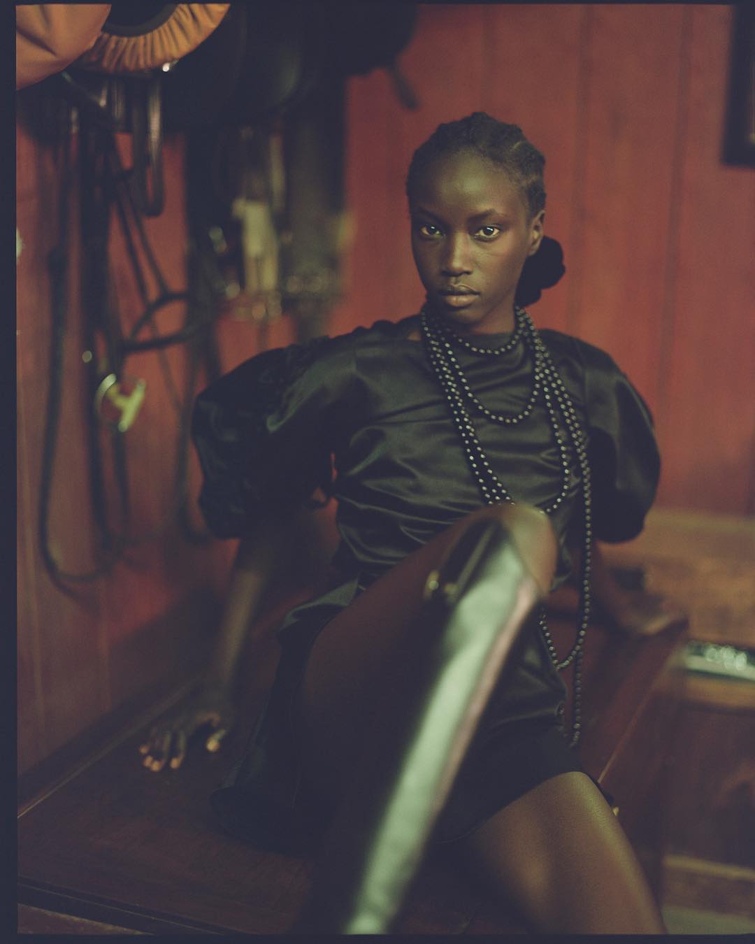 Elegant dark-skinned woman in black dress and leather boots sits on dark surface with horse riding equipment in background.